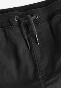 Pantalon de survêtement noir avec une taille côtelée, une cordelière et des œillets en métal. Le tissu a une texture lisse et des détails de couture minimaux.