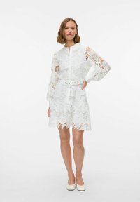 Weißes Spitzenkleid mit langen Ärmeln, floralem Muster, kragenförmigem Ausschnitt und einer taillierten Taille mit einem Gürtel. Der Saum ist gewellt.