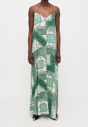 Frau trägt ein weißes Maxikleid mit grünen Paisley- und Blumen-Patchworkmustern, dünnen Trägern und schwarzen spitzen Pumps.