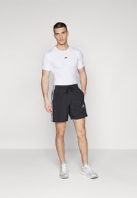 Hvid tætsiddende t-shirt med sort logo, parret med sorte træningsshorts med hvide striber og et logo. Grå træningssko.