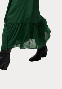 Lång grön klänning med en utsvängd nederkant, tillverkad av lättviktsmaterial, i kombination med svarta vadboots med klack.