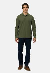 Hosszú ujjú, olíva zöld pólóing, gallérral, sötétkék farmernadrággal és barna, fűzős csizmával párosítva. Egyszerű dizájn, látható minták nélkül.