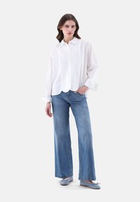 Weißes Hemd mit Rundhalsausschnitt und gewelltem Saum, kombiniert mit hellblauen weit geschnittenen Jeans und blauen Ballerinas, vor einem neutralen Hintergrund.