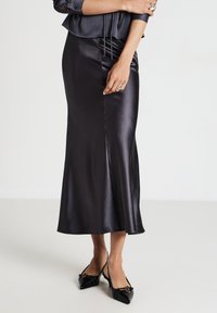 Donna che indossa una gonna midi di satin nera e tacchi a punta neri, con le braccia incrociate e anelli sulle dita visibili.