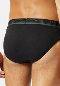 Ein Mann trägt schwarze Slips mit einem elastischen Bund, auf dem das Wort "uncover" auf einem hellen Hintergrund zu sehen ist.