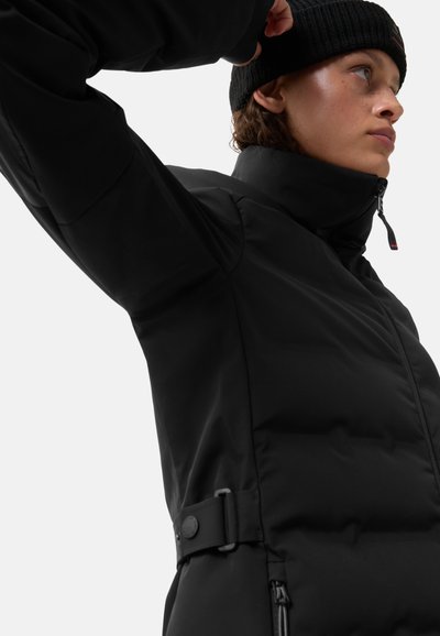 Zwarte gewatteerde jas met hoge kraag, ritssluiting en ceintuurdetail in de taille. Gladde stof en getailleerd ontwerp. Zwart gebreide muts gedragen.