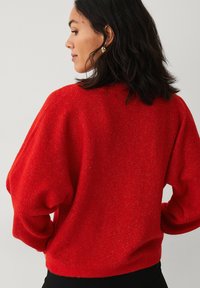 Rød strikket sweater med afslappet pasform, lange ærmer og ribbet halsudskæring. Har en subtil tekstur og en blød finish, parret med sorte bukser.