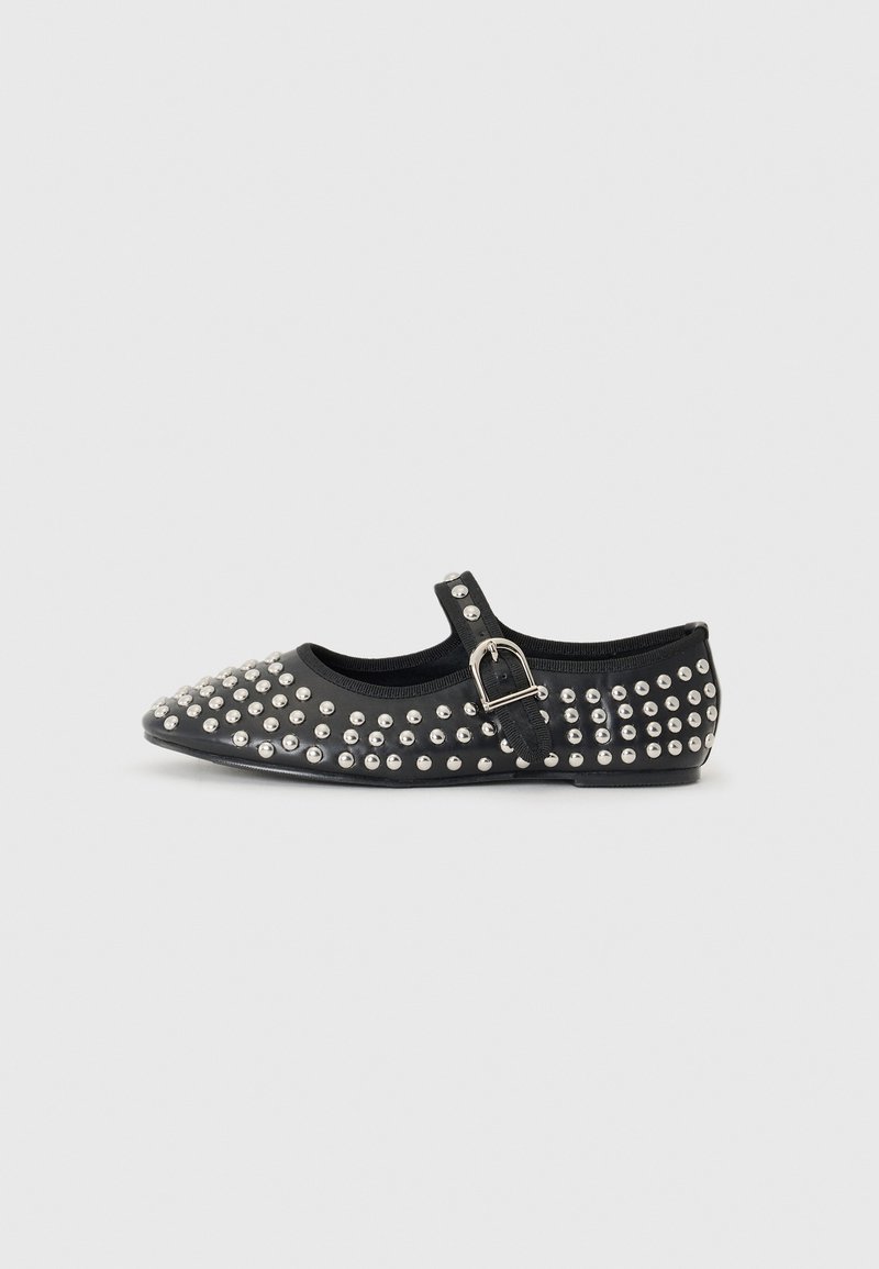 Chaussure plate noire de type Mary Jane avec des clous argentés et une bride à boucle ajustable sur un fond clair uni.