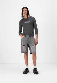 Chemise gris à manches longues avec logo, associée à un short en denim gris délavé. Le modèle porte des chaussures noires épaisses et des chaussettes blanches. Ensemble simple et décontracté.