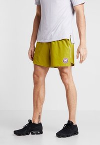 Grüne Sportshorts mit einem Logo-Patch, seitlicher Reißverschlusstasche, leichtem Stoff, kombiniert mit einem grauen kurzärmeligen Shirt und schwarzen Turnschuhen.