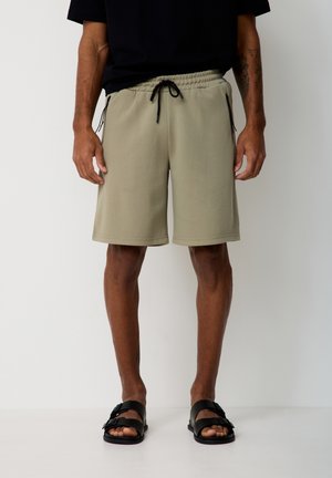 Mann trägt beige Shorts mit Kordelzug und seitlichen Reißverschlusstaschen sowie schwarze Sandalen, steht vor einfarbigem weißem Hintergrund.