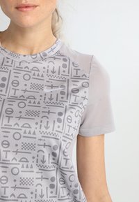 Šedé sportovní tričko s texturovanou látkou a grafickým vzorem geometrických tvarů a symbolů, doplněné síťovanými detaily.