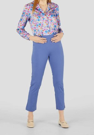 Kvinde iført en farverig blomsterbluse stoppet ned i højtaljede, slim fit blå bukser med cremefarvede loafers på en lys grå baggrund.