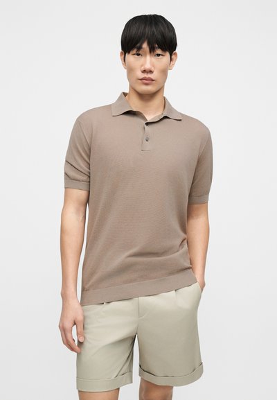 Πόλο με κοντά μανίκια σε πλεκτό ύφασμα ανοιχτού καφέ χρώματος, με κλασικό γιακά, πατιλέτα με δύο κουμπιά και άνετη εφαρμογή. Συνδυάζεται με μπεζ σορτς.