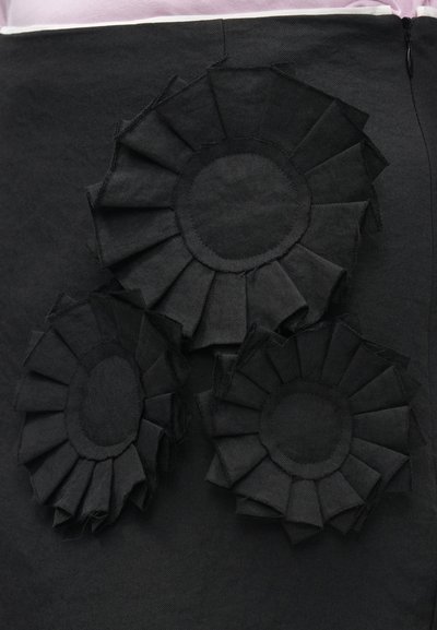 Μαύρη φούστα από ύφασμα με τρία εμφανή υφασμάτινα μαύρα τριαντάφυλλα με στρωματοποιημένες πιέτες. Η φούστα έχει λείο φινίρισμα και κρυφό φερμουάρ.