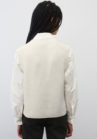 Cremfarbener, gestrickter ärmelloser Pullover mit strukturierter Oberfläche. Gerippter Saum und Schulternähte. Über einem weißen Hemd mit Kragen getragen.