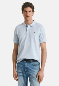 Jonge man met krullend haar, gekleed in een lichtblauw polo-shirt en blauwe jeans, staand met ontspannen handen langs zijn zij.