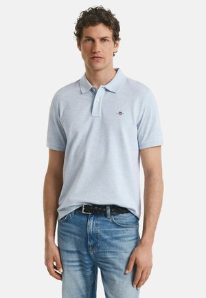 Jeune homme avec des cheveux bouclés portant un polo bleu clair et un jean bleu, se tenant avec les mains détendues sur les côtés.