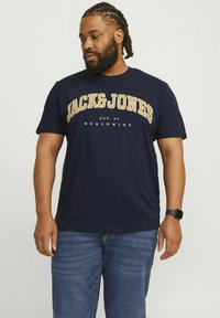 Námornícky modré bavlnené tričko s bielym a žltým textom loga "JACK&JONES" a nápisom "EST. 90 WORLDWIDE" pod ním. Klasický dizajn s okrúhlym výstrihom.