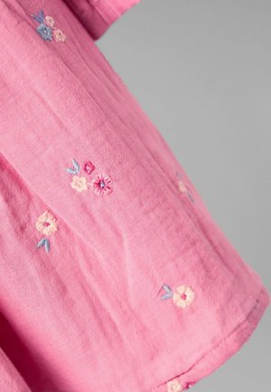 Chemise rose en tissu doux, ornée de délicbroderies florales délicates dans des couleurs pastel. Les accents incluent des nuances de bleu clair et de crème.
