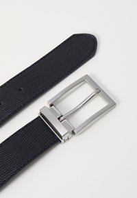 Zwarte leren riem met textuur, voorzien van een rechthoekige zilveren metalen gesp en meerdere gaatjes voor maatverstelling. Gladde afwerking en slank ontwerp.