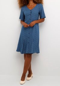 Kvinna i en knälång blå blommig klänning med knappar och korta ärmar, kombinerad med krämfärgade låga klackskor.