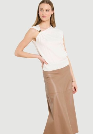 Naine, kes kannab valget varrukateta topi metallist rõnga detailiga ja beežist nahast kõrge vöökohaga midi-seelikut, poseerib ühe käega puusal.