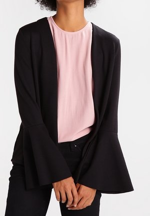 Woman wearing a pale pink blouse and black cardigan with wide bell sleeves, standing with hands loosely clasped in front.