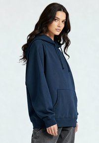 Jeune femme aux longs cheveux noirs portant un sweat à capuche bleu marine et un pantalon gris, debout sur un fond clair uni, légèrement de profil.