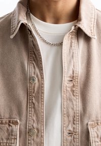 Lys beige denimjakke med trykknapper og forlommer, båret over en hvid t-shirt, der har en sølv kædND.