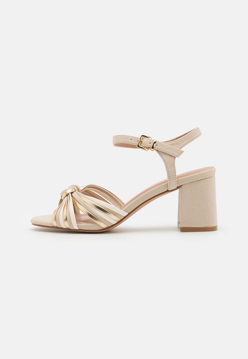 Sandale à talon en bloc beige avec un design de devant noué, des brides métalliques dorées, une bride de cheville et un dessus en texture douce.