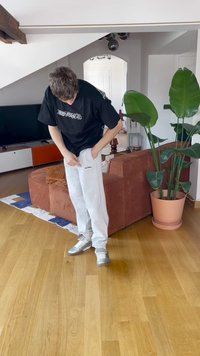 Pantalones de chándal grises con detalles de texto, camiseta negra de gran tamaño y zapatillas plateadas; en interiores con una planta y una alfombra de estampado en el fondo.