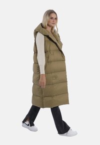 Olivgrüner Steppwesten mit Kapuze, die große, gesteppten Abschnitte und Druckknöpfe aufweist. Das Modell trägt sie über einem cremfarbenen Oberteil und schwarzen Jeans.