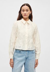 Kremowa, fakturowana koszula na guziki z kołnierzykiem i długimi rękawami, zestawiona z jasnoniebieskimi dżinsami. Posiada subtelne kwiatowe wzory.