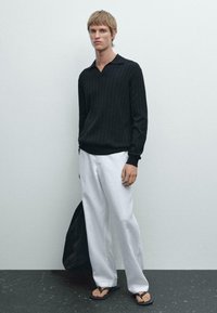 Zwarte geribbelde poloshirt met lange mouwen, gecombineerd met losse witte broek en slippers. Houdt een zwarte tote bag vast tegen een effen achtergrond.