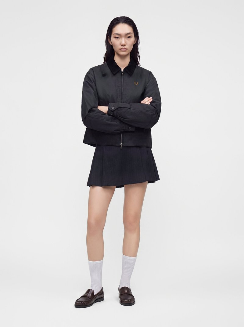 Le modèle se tient debout, les bras croisés, vêtu d’une veste noire zippée et d’une jupe plissée, avec des chaussettes blanches et des mocassins foncés, sur un fond blanc uni.