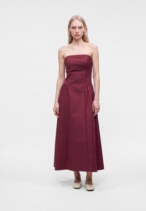 Vrouw met lang blond haar, gekleed in een strapless bordeaux midi-jurk en beige schoenen, staand tegen een effen lichte achtergrond.