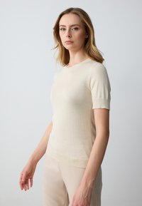 Maglione beige a maniche corte in morbido materiale a maglia, con scollatura rotonda, caratterizzato da un design aderente e sottili dettagli a costine su maniche e orlo.