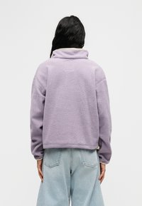 Μωβ φλις πουλόβερ με ψηλό γιακά, χαμηλωμένους ώμους και απαλό υλικό. Διαθέτει κίτρινη λεπτομέρεια κατά μήκος του γιακά και των μανικιών.