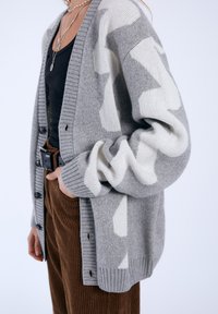 Gray and cream knit cardigan with a geometric pattern, ribbed cuffs, and buttons. Worn over a black top and paired with brown corduroy pants.