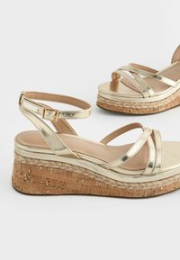 Goldene metallische Keilsandalen mit gekreuzten Riemen, Korktextur-Sohle und glattem, gepolstertem Fußbett. Verstellbarer Knöchelriemen vorhanden.
