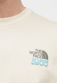 Κοντινό πλάνο πουκάμισου εκρού με το λογότυπο "The North Face 1966" σε μαύρο και ανοιχτό γαλάζιο στο στήθος.