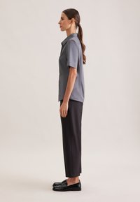 Camicia grigia a maniche corte con bottoni e colletto, abbinata a pantaloni neri a gamba stretta e mocassini neri lucidi, vista laterale.