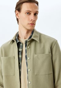 Giacca camicia verde oliva con ampie tasche frontali, chiusura a bottoni e tessuto texture. Indossata sopra una camicia di flanella a quadri e un maglione beige.