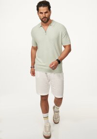 Hellgrünes, strukturiertes Poloshirt mit Reißverschlusskragen, kombiniert mit weißen, lässigen Shorts und beigen Sneakern mit gelben Akzenten.