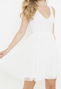 Nainen, joka pitää yllään hihattoman valkoisen mekon, jossa on paljeteilla koristeltu yläosa ja laskostettu tiulihame, venyttää hametta hieman sivulle.