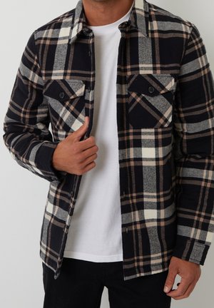 Mand iført sort, gråt og beige ternet skjorte over en almindelig hvid t-shirt, justerer skjorten med sin venstre hånd.