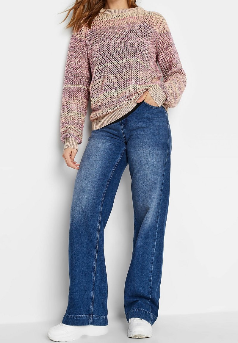 Suéter de punto multicolor con mangas largas y puños acanalados, combinado con unos vaqueros de mezclilla azules de pierna ancha y cintura alta. Zapatillas blancas completan el look.