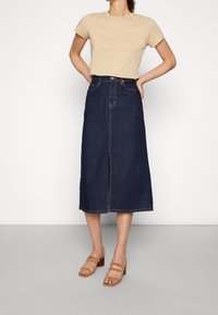 Mörkblå jeansmidirandig kjol med slits framtill, hög midja och kontrasterande sömmar. Kombinerad med en beige kortärmad topp och ljusbruna sandaler.