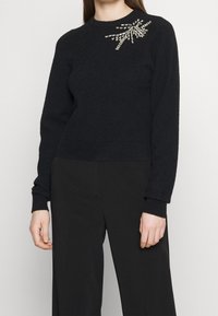 Czarny sweter z okrągłym dekoltem, długimi rękawami i teksturowanym materiałem, z dekoracyjnymi srebrnymi ozdobami na jednym ramieniu.